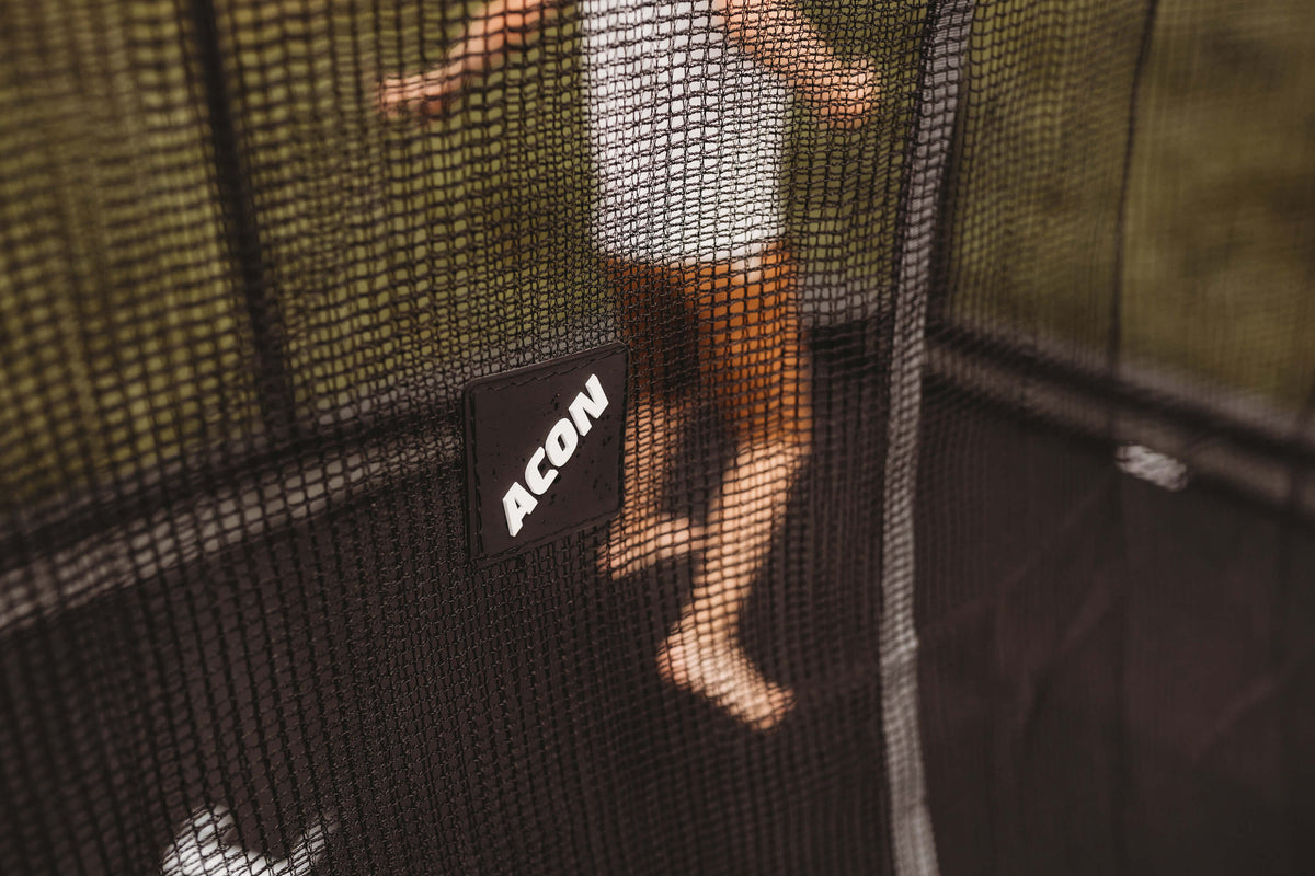 A kid jumping on an ACON trampoline with enclosure. The enclosure has a tightly-sewn mesh and a logo of the trampoline manufacturer Acon.