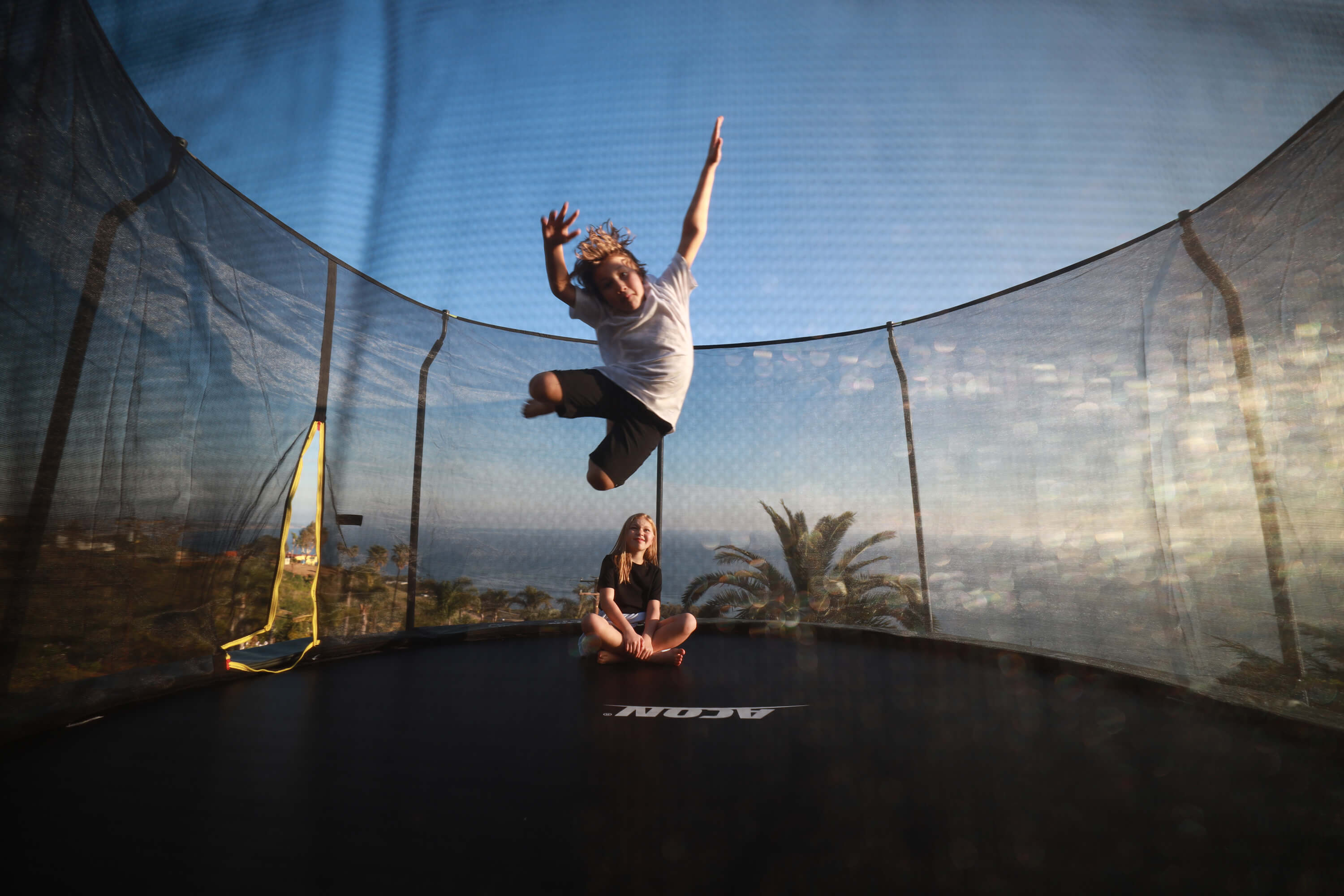 A boy jumps on a round trampoline, a girl sits on the edge and watches.