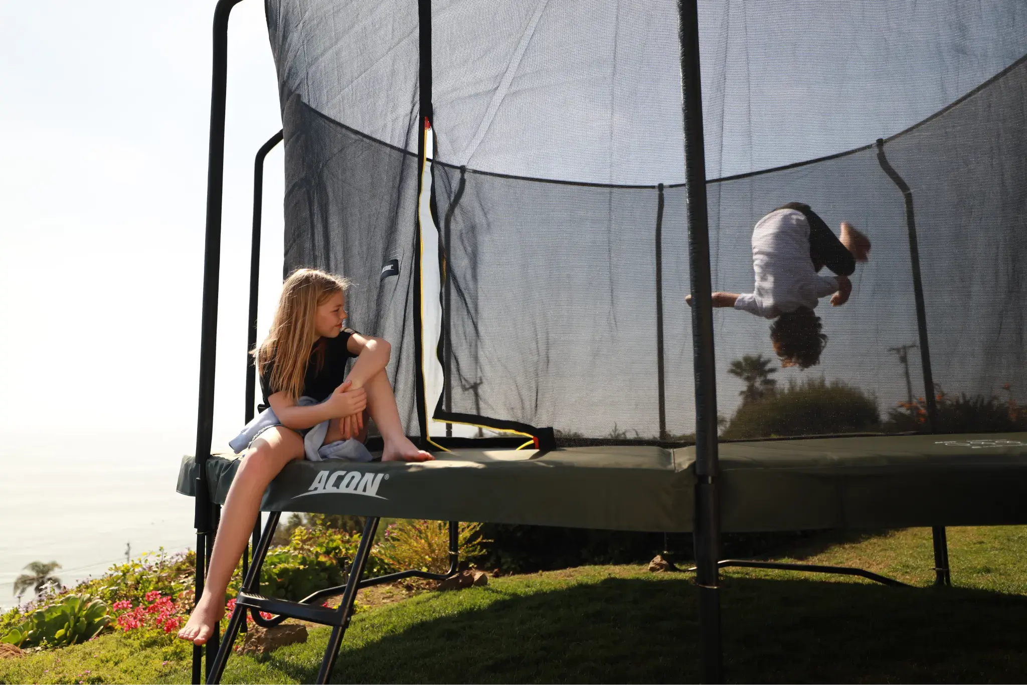 A child performs a backflip on an Acon trampoline with a safety net, while another child sits on the trampoline edge, watching.