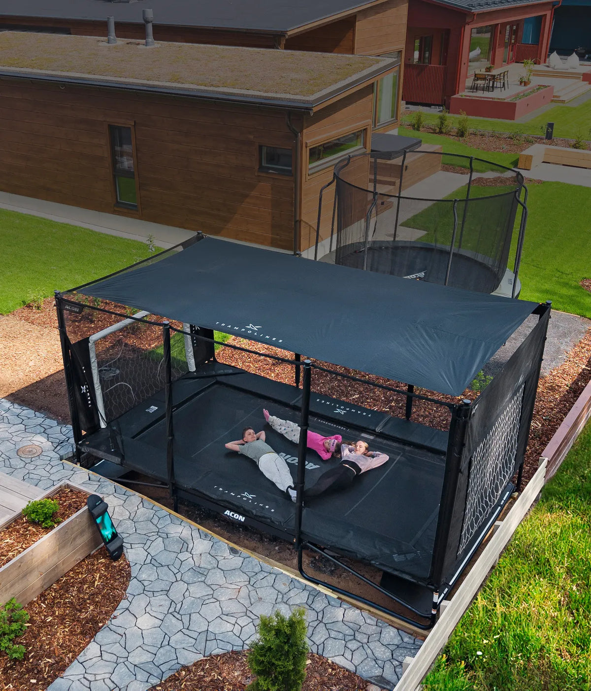 Children lying on an Acon X trampoline under the Sun Shade, relaxing and resting together, showing shaded backyard play and UV protection.
