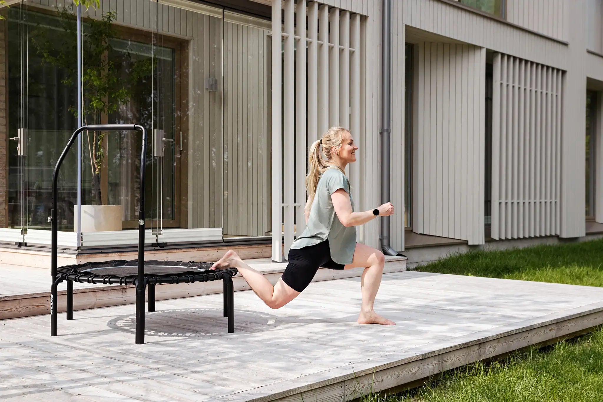 Woman with blonde hair doing a split squat on an Acon rebounder trampoline in a sunny outdoor setting.