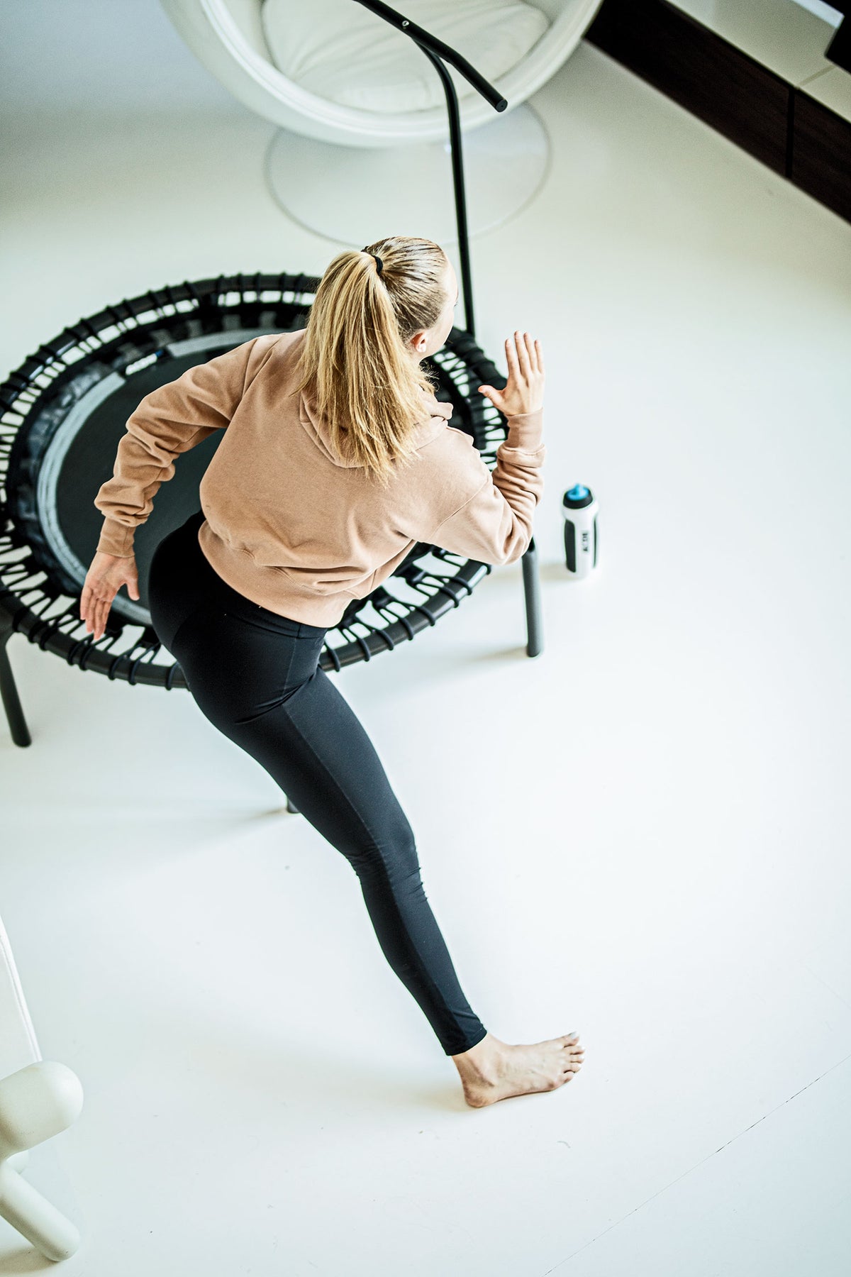 A woman exercising barefoot on an Acon fitness trampoline with a handlebar in a bright white studio.
