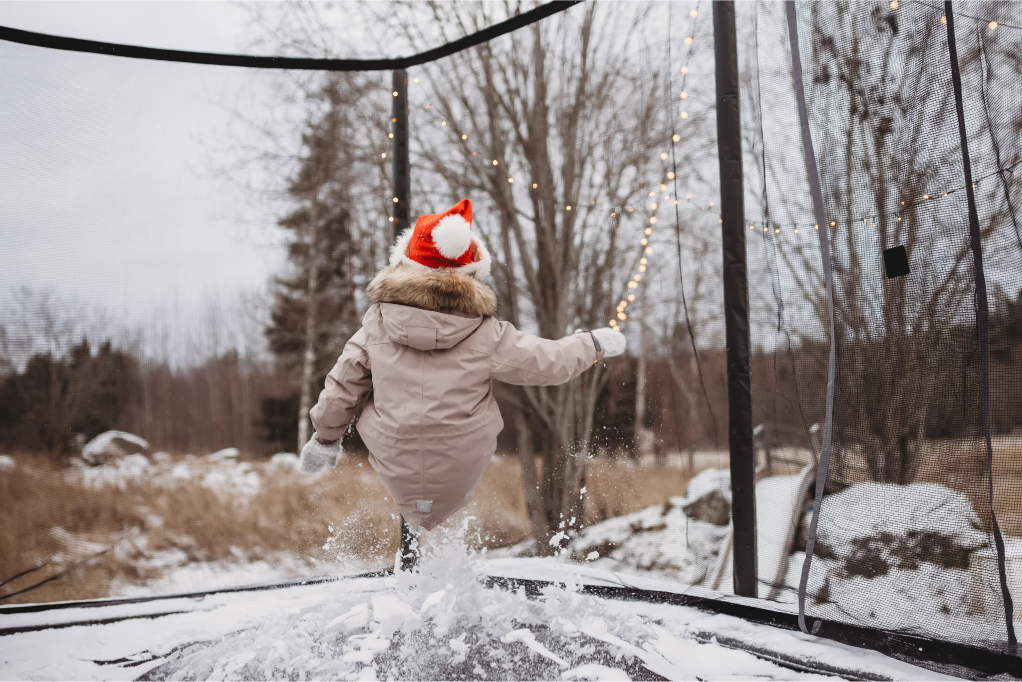 Child with a winter coat and a santa hat jumping on a trampoline with snow.