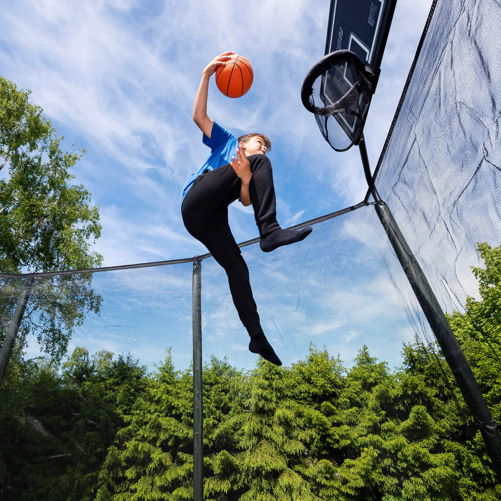 A boy jumps holding his leg and throws a basketball into the trampoline basketball hoop.