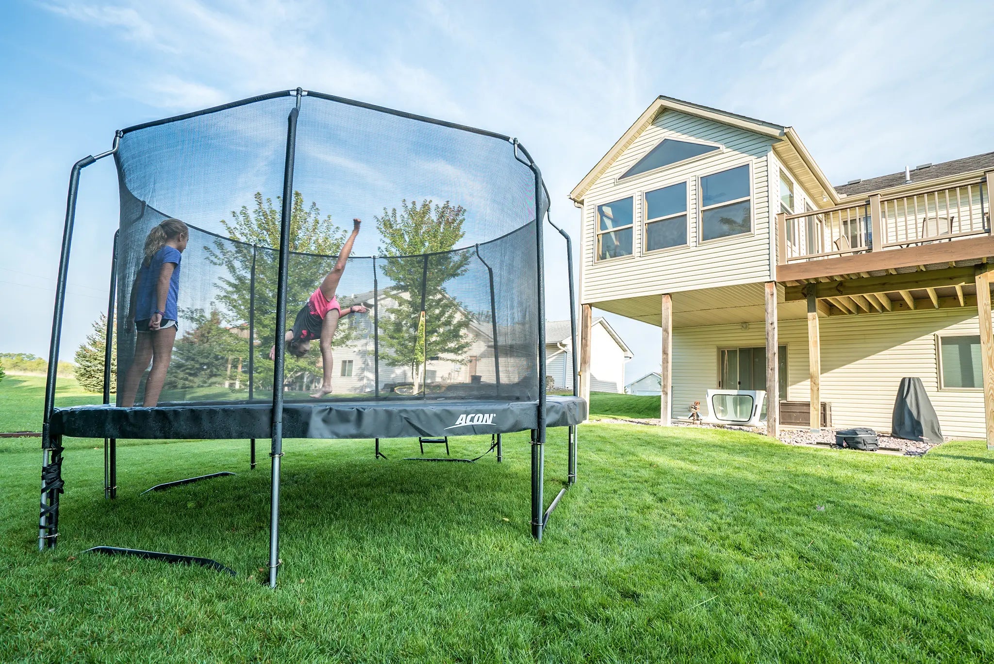 Two girls on a Acon trampoline, other is performing a trick while the other watches. 