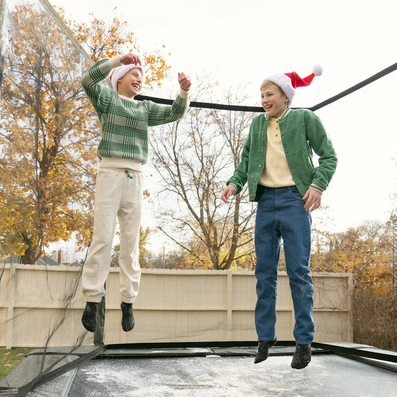 Two kids wearing Santa hats laughing and jumping on a trampoline with light snow.
