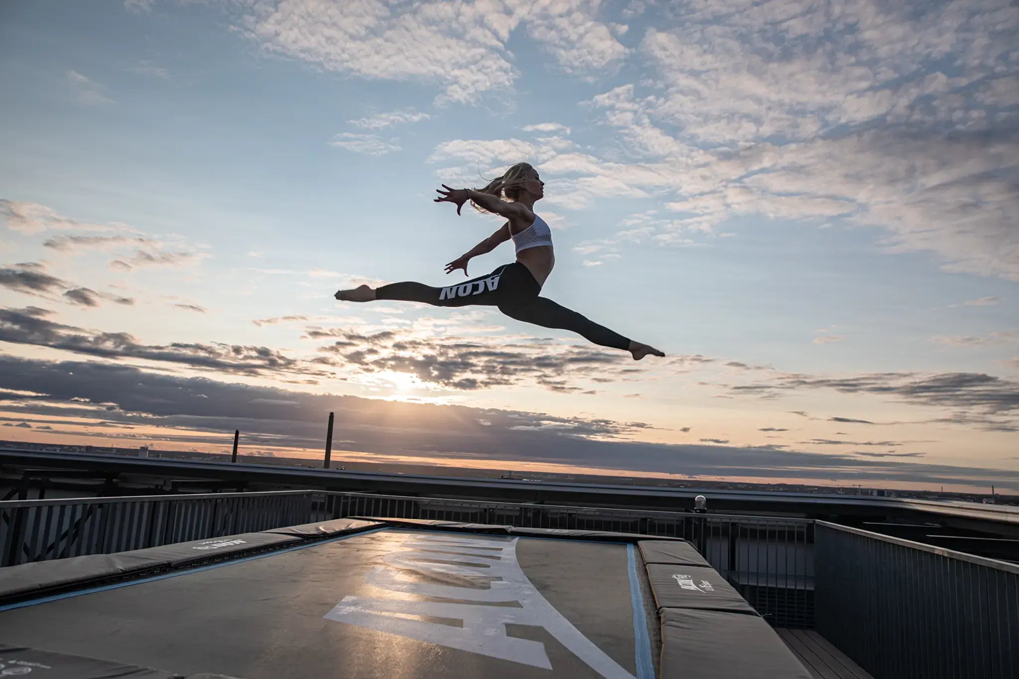 Woman performing a split jump on an Acon trampoline at sunset on a rooftop.