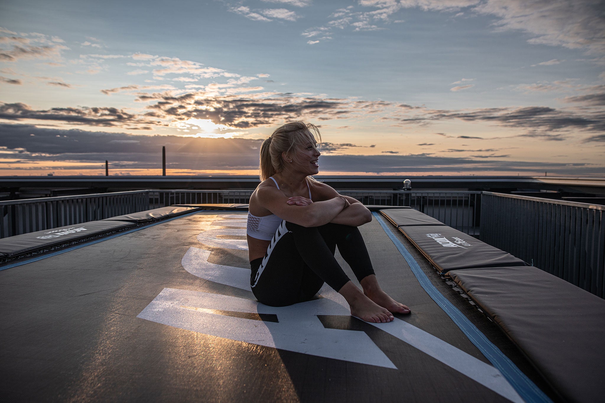 Cheerful woman sitting on an Acon 16 HD trampoline with a sunset as background