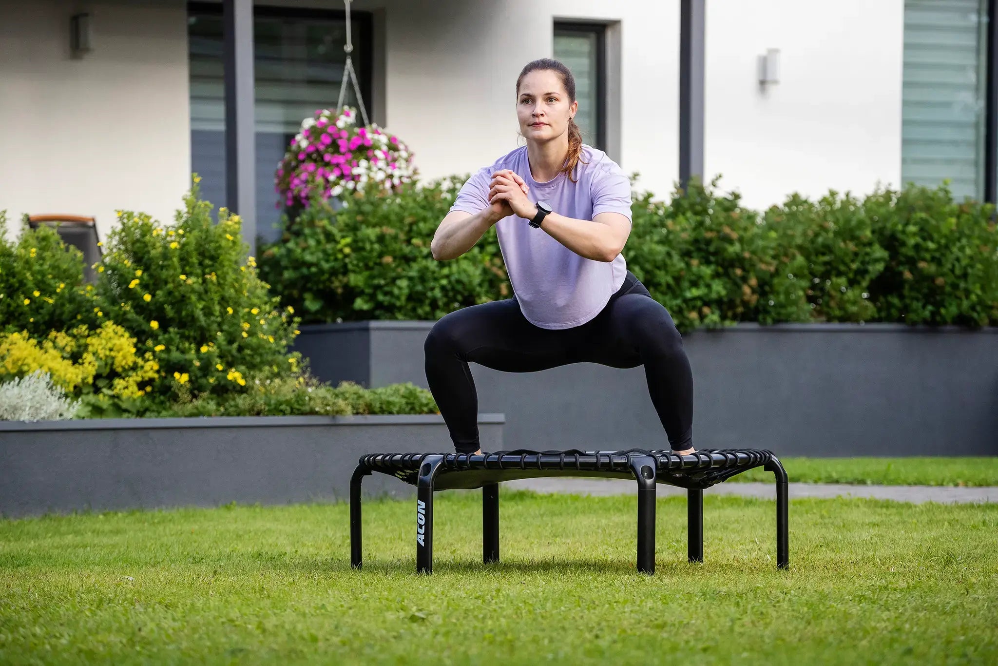 Woman doing a squat exercise on an Acon fitness trampoline in a garden setting.